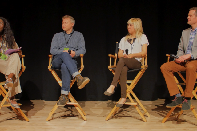 4 people sit in directors chair on a stage, a woman on the far left holds a microphone, a man in a blue shirt, a woman in a white shirt, and very tall man in gray jacket from right to left